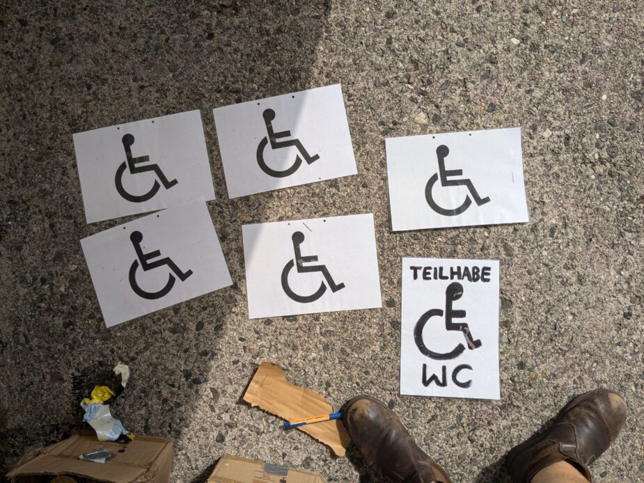 Auf dem Boden liegen 6 A4 Blätter, auf denen Piktogramme mit einem Mensch im Rollstuhl gemalt sind. Auf einem steht zusätzlich "Teilhabe WC" geschrieben