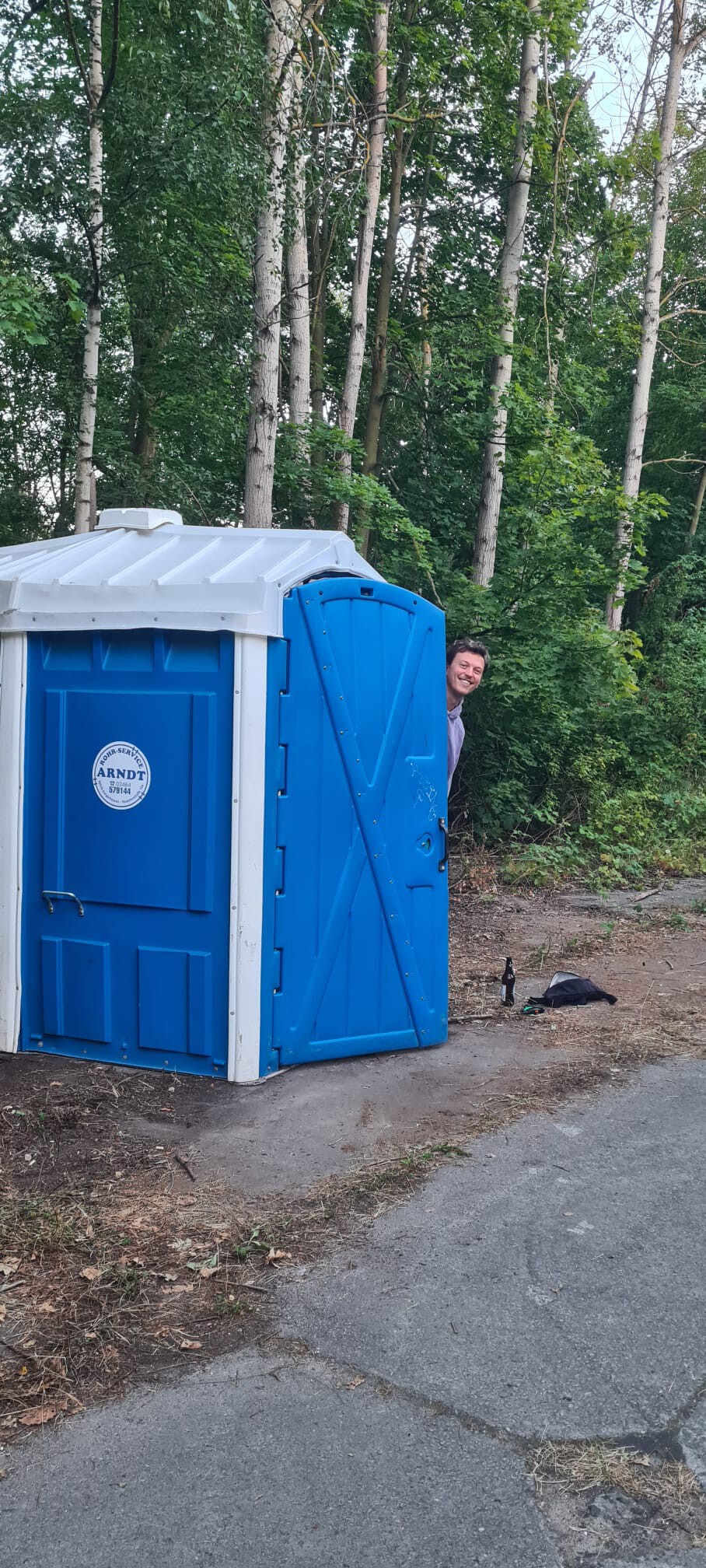 Ein großes blaues barriere-frei Dixie. Die Tür ist offen, eine Person guckt um die Ecke heraus. Im Hintergrund sind Birken.