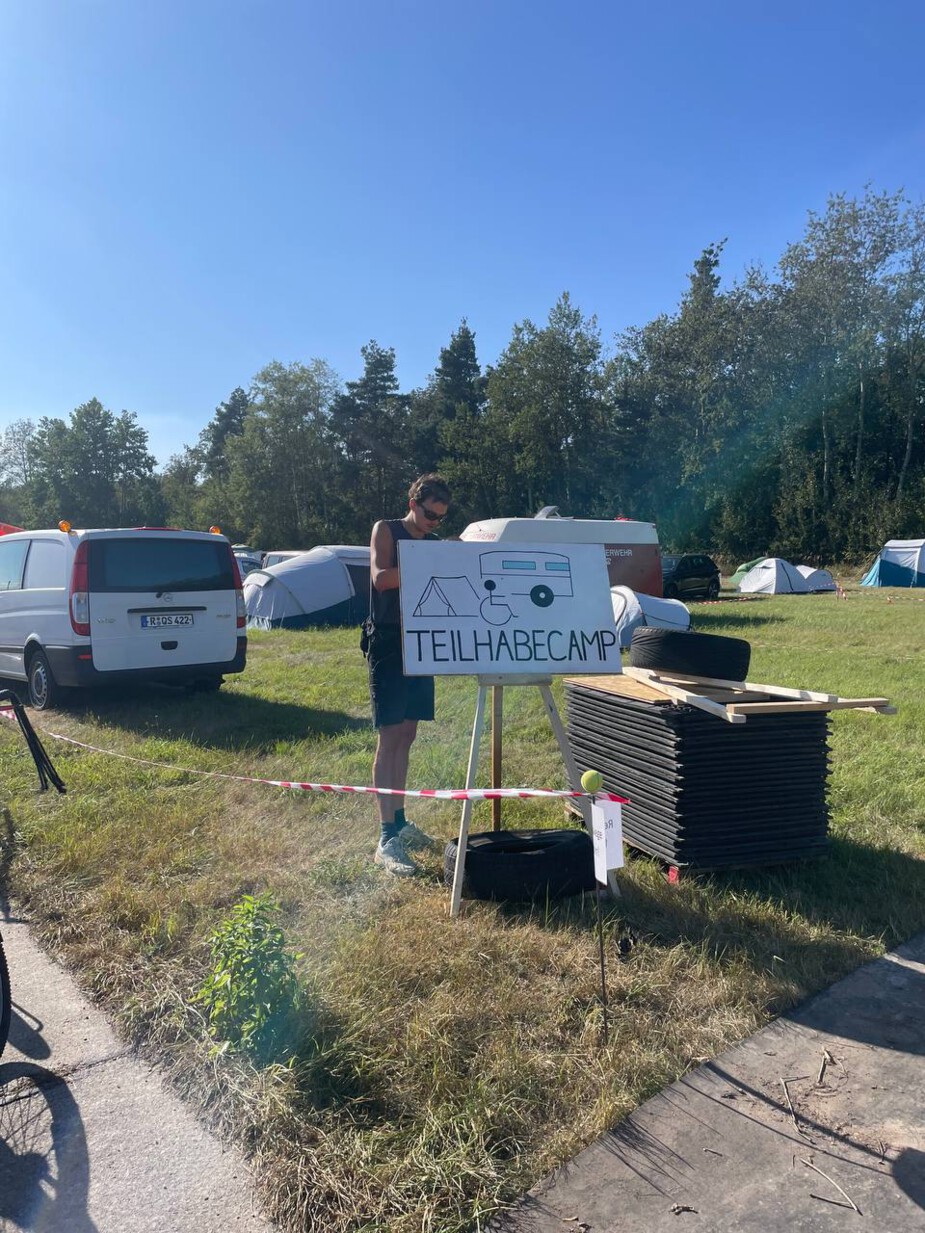 Eine Person stellt ein Schild in einem abgeflatterten Bereich, mit der Aufschrift Teilhabe Camp auf. Rechts von ihr stehen viele schwarze Schwerlastmatten auf einer Europ-Palette. Die Person steht auf einer Camping-Wiese, weshalb im Hintergrund PKWs und Zelte zu sehen sind.