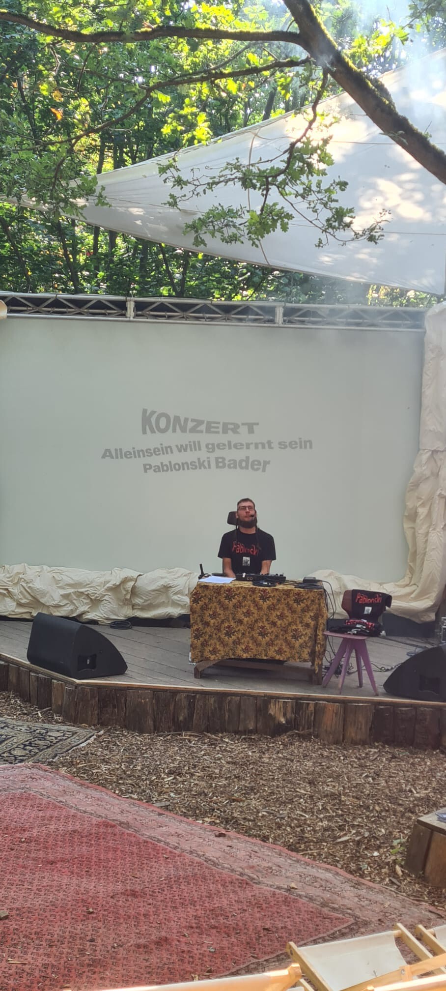 Auf einer Bühne vor einer großen Leinwand sitzt Künstler Pablonski. Er sitzt mit seinem E-Rollstuhl vor einem Tisch, auf der eine Loop-Station steht. Auf der Leinwand steht: "Konzert. Alleinsein will gelernt sein. Pablonski Bader."